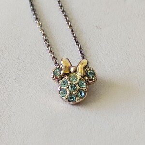 May include: A gold-toned metal necklace featuring a pendant shaped like a mouse head with a bow. The pendant is embellished with multiple light blue circular crystals set into the metal surface, suspended from a delicate matching metal chain.