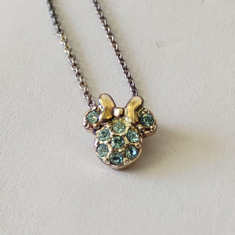 May include: A gold-toned metal necklace featuring a pendant shaped like a mouse head with a bow. The pendant is embellished with multiple light blue circular crystals set into the metal surface, suspended from a delicate matching metal chain.