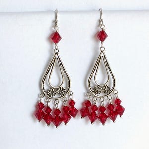 May include: A pair of silver-toned dangle earrings featuring a teardrop-shaped frame with twisted wire detailing. Each earring is accented with a single red bicone crystal at the top and five smaller red bicone crystals hanging from the base.