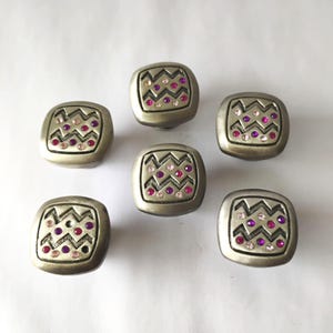 May include: Six square metal knobs with rounded corners, featuring a matte silver finish. Each knob has a central recessed area decorated with a zigzag pattern and embedded small circular crystals in shades of pink, purple, and clear white.