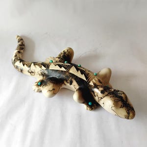 May include: A ceramic lizard sculpture with a cream-colored body, adorned with black patterns and turquoise accents. The lizard features a detailed design with geometric shapes and is a decorative art piece.