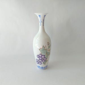 May include: A white ceramic vase with a slender neck and flared top. The vase features a painted design of purple grapes, green leaves, and white flowers. Blue decorative patterns adorn the top and base of the vase. The vase is approximately 12 inches tall.