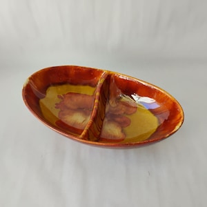 May include: An oval-shaped ceramic serving dish featuring a central divider. The piece has a glossy glaze with a color palette of deep burnt orange and mustard yellow, displaying a blended, marbled pattern throughout the interior surface.