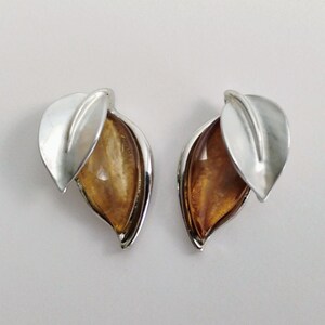 May include: A pair of silver-toned earrings featuring a polished metal leaf design overlapping a teardrop-shaped amber-colored stone. The earrings have a smooth, metallic finish and a pointed, organic shape that mimics the appearance of a leaf.