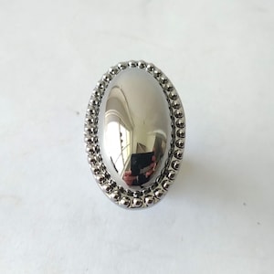 May include: Oval-shaped, silver-toned metal knob with a beaded border. The center of the knob is smooth and reflective, showing a distorted reflection. The knob is likely for furniture or decorative use.