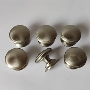 May include: A set of six round cabinet knobs with a brushed silver metallic finish. Five of the knobs are positioned with their flat circular faces pointing upward, while one knob is placed on its side to show the stem and circular base.