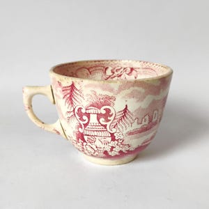 May include: A ceramic teacup featuring a cream-colored base with a detailed red transferware pattern. The design depicts a central decorative urn surrounded by floral elements, trees, and architectural structures, with a matching red rim pattern.