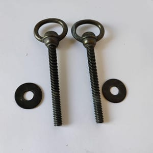 May include: Two antique brass-toned metal eye bolts with threaded shafts and circular heads, accompanied by two separate flat metal washers. The hardware features a weathered, dark metallic finish with visible threading along the vertical stems.
