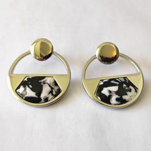 May include: A pair of geometric earrings. Each earring features a gold-colored stud, a silver-colored circular frame, and a gold-colored, half-hexagon shape with a black and white marbled design.