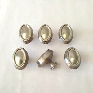 May include: A set of six oval-shaped cabinet knobs with a smooth, brushed metallic finish. Five of the knobs are displayed from a front-facing perspective, while one central knob is positioned at an angle to reveal its stem and mounting base.