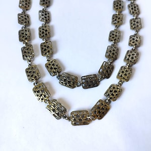 May include: A long necklace composed of two strands of interconnected rectangular metal links. Each link features an openwork, textured, grid-like pattern in a muted, antique brass tone. The chain is constructed with small circular jump rings.