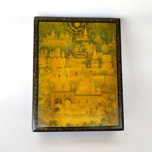 May include: A rectangular, hand-painted box with a detailed landscape scene. The artwork depicts a golden-hued cityscape with buildings, towers, and figures, framed by a black border with gold filigree. The box is set against a white background.