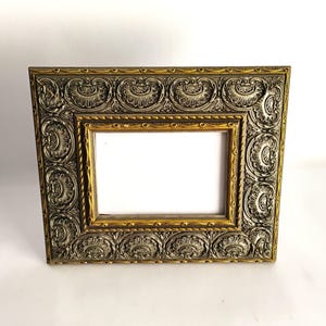 May include: A rectangular picture frame featuring a metallic silver finish with raised, repeating shell-like motifs along the border. The inner edge of the frame is accented with a thin, gold-toned trim, surrounding a plain white rectangular center.