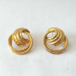 May include: A pair of gold-colored stud earrings. Each earring features a textured, rope-like outer ring and a central, layered design. The earrings have a post backing for pierced ears and are set against a white background.