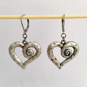 May include: A pair of silver-toned heart-shaped earrings. Each earring features a hammered texture and a spiral design within the heart. The earrings hang from lever-back closures.