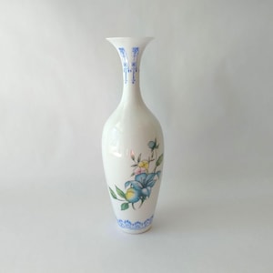 May include: A white ceramic vase with a slender neck and flared opening. The vase features a floral design with blue, yellow, and green accents. Blue decorative patterns adorn the neck and base, creating an elegant decorative piece.