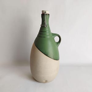 May include: A ceramic bottle with a rounded base and a narrow neck, featuring a two-tone design with a matte cream-colored lower section and a smooth forest green upper section. It includes a side handle and a metal swing-top stopper mechanism.