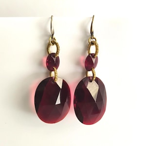 May include: A pair of dangle earrings featuring faceted, oval-shaped, deep red gemstones. The earrings have gold-toned metal accents and ear wires. The design includes smaller, oval-shaped gems above the larger ones, connected by gold-toned rings.