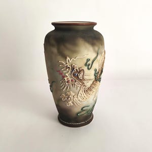 May include: A ceramic vase with a matte, smoky gray and brown gradient finish. The exterior features a raised, textured relief of a dragon in cream, green, and red tones. The rim and base are dark brown, with the base accented by a row of white dots.