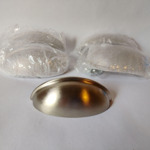 May include: A brushed nickel finish cup pull drawer handle with a smooth, semi-circular shape. The handle features a curved front surface and two mounting holes on the flat base. Two additional identical handles are visible in clear plastic packaging.