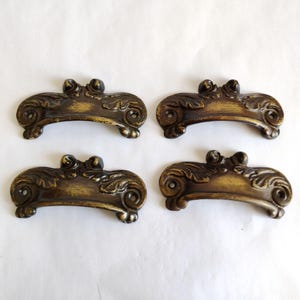 May include: Four antique bronze-colored drawer pulls with ornate, scroll-like designs. Each pull features a curved handle with decorative floral elements and two screw holes for mounting. The pulls have a distressed finish.