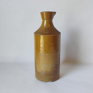 May include: A cylindrical brown stoneware bottle with a narrow neck and a flared rim. The surface features a glossy glaze and embossed text near the base that reads F. & J. ARNOLD LONDON ENGLAND, with additional faint lettering above.