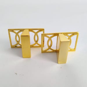 May include: Two gold-colored metal cabinet pulls featuring a rectangular frame with an openwork design of interlocking circles. Each pull has a solid, smooth, vertical rectangular handle attached to the center of the decorative frame.