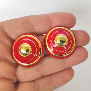 May include: A pair of round, button-style earrings featuring a vibrant red enamel finish with concentric gold-tone metal rings and a polished gold-tone center dome. The design consists of layered circular elements that create a tiered, dimensional appearance.