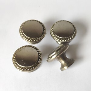 May include: Four circular cabinet knobs with a metallic silver finish. Each knob features a smooth, flat center disc surrounded by a raised, twisted rope-style border. The knobs have a tapered base and a polished, reflective surface texture.