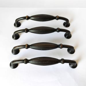 May include: Four dark bronze cabinet pulls arranged in a vertical row. Each handle features a central, elongated, ribbed oval section flanked by two smooth, curved arms that terminate in circular mounting bases with a weathered, metallic finish.