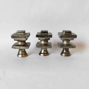 May include: Three silver-toned metal finials, each featuring a tiered square design with a central pedestal base. The metallic surface has a textured, weathered finish with visible highlights and shadows on the stacked geometric shapes.