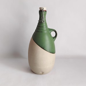 May include: A two-toned ceramic bottle with a green top and a cream-colored base. The bottle has a handle and a metal clasp with a cork stopper. The design is simple and elegant, suitable for storing liquids.