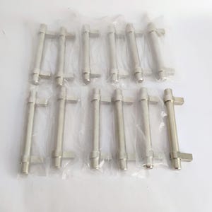 May include: A collection of ten brushed nickel cabinet pulls, each individually wrapped in clear plastic. The pulls have a cylindrical body with a rectangular handle. The hardware is designed for use on drawers and cabinets.