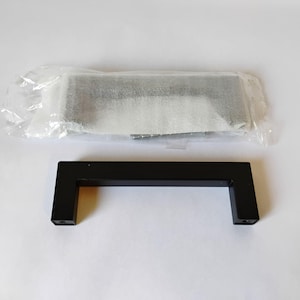 May include: A matte black cabinet handle with a rectangular, U-shaped design. The handle features two mounting points at the ends. A second identical handle is visible above it, enclosed in a clear plastic protective bag with included screws.