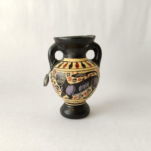 May include: A small, black and yellow ceramic vase with two handles. The vase features a painted design of a mythical creature in black, purple, and red. A small metal charm hangs from one handle. The vase has a round base.