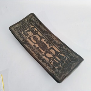 May include: A rectangular, dark brown wooden tray with a carved design. The design features a central figure surrounded by floral and geometric patterns. The tray is likely handmade and has a rustic aesthetic.