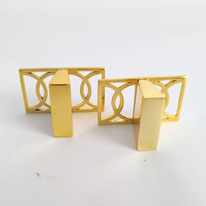 May include: Two gold-colored cabinet pulls. Each pull has a rectangular backplate with a geometric design of interlocking circles. A solid, rectangular handle extends from the backplate. The pulls are made of metal and are set against a white background.