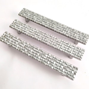 May include: Three silver cabinet pulls with a textured, square pattern. Each pull has a rectangular bar with a series of small squares and two rounded posts for mounting. The hardware is displayed on a white surface.