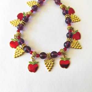 May include: A charm bracelet featuring translucent purple beads and small red beads, accented with gold-toned metal charms shaped like red apples and clusters of grapes, all connected by small gold-toned spacers along the circular strand.