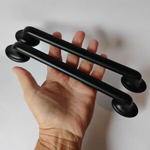 May include: Two matte black cabinet pulls with a smooth, cylindrical bar design and rounded mounting bases at each end. These hardware pieces feature a solid, uniform dark finish and a simple, functional shape for furniture or door installation.