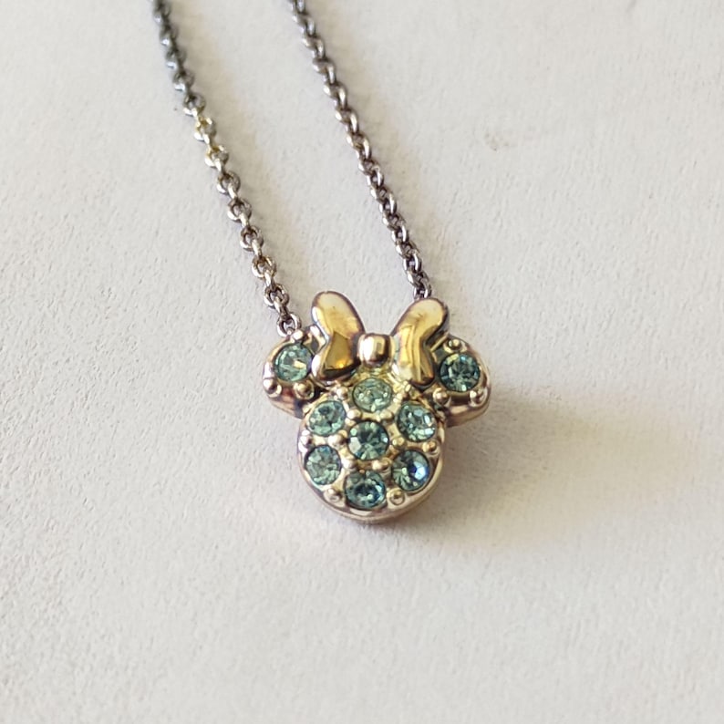May include: A gold-toned metal necklace featuring a pendant shaped like a mouse head with a bow. The pendant is embellished with multiple small, light blue circular crystals set into the metal surface, suspended from a delicate link chain.