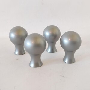 May include: A set of four silver-colored cabinet knobs, each featuring a smooth, spherical top and a tapered base. These hardware pieces have a uniform metallic finish and a rounded shape, designed for attachment to furniture or cabinetry doors.
