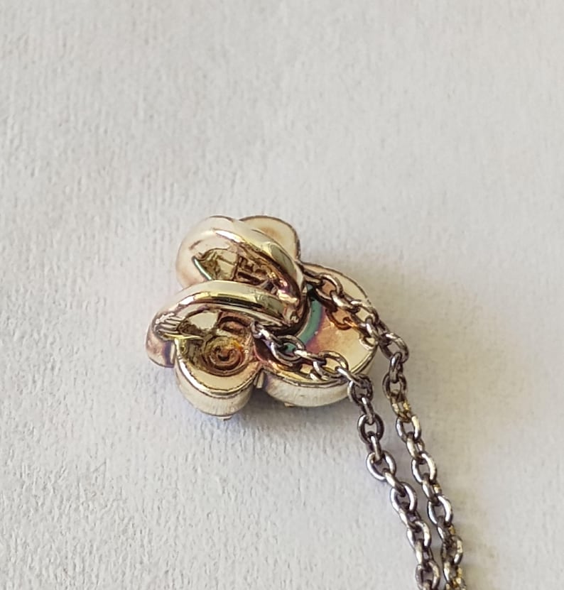 May include: A gold-toned metal pendant shaped like a three-petaled flower or clover, featuring a central circular detail and a polished finish. The pendant is attached to a delicate, thin metal chain link necklace that extends downward from the piece.