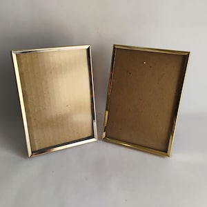 May include: Two rectangular gold-colored picture frames. The frames have a shiny, metallic finish and a simple, elegant design. The interior of each frame is a light brown color, ready for a photo or artwork. The frames are displayed on a white surface.