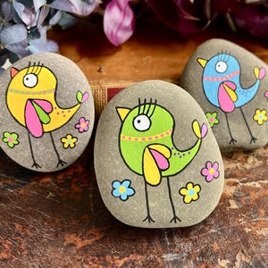 Puede incluir: Cuatro piedras pintadas con diseños de pájaros coloridos. Cada pájaro está pintado con colores brillantes como amarillo, azul, rosa y verde, y está rodeado de pequeños acentos florales. Las piedras están sobre una superficie de madera.