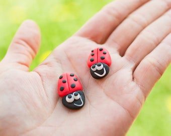 May include: Two red and black ladybug figurines with white eyes and smiling mouths, held in an open hand against a green background.