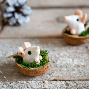 Può includere: Due figurine di topo bianco sono annidati in gusci di noce, ognuno con un letto di muschio verde. I gusci sono seduti su una superficie di legno marrone chiaro.