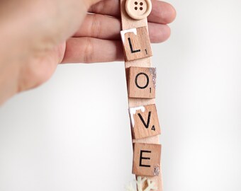 May include: A wooden ornament featuring the word "LOVE" spelled out using Scrabble tiles. The ornament is decorated with a white snowflake and a wooden button.