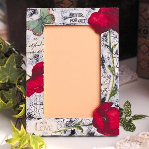 Puede incluir: Marco de fotos rectangular con un acabado blanco y negro envejecido, adornado con amapolas rojas y una mariposa verde. El marco tiene las palabras "NEVER FORGET" y "WITH LOVE". El marco está rodeado de vegetación.