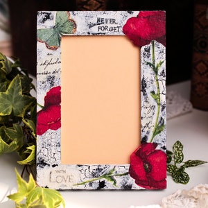 Pode incluir: Um quadro de fotos branco com acabamento envelhecido. O quadro é decorado com papoulas vermelhas, folhas verdes e uma borboleta. O quadro tem o texto "Never Forget" e "With Love" impresso nele.